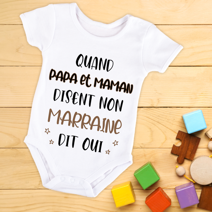 Body bébé annonce: body blanc pour annonce grossesse, texte humoristique "Quand papa et maman disent non marrainne dit oui" avec blocs de couleur et jouets en bois autour.