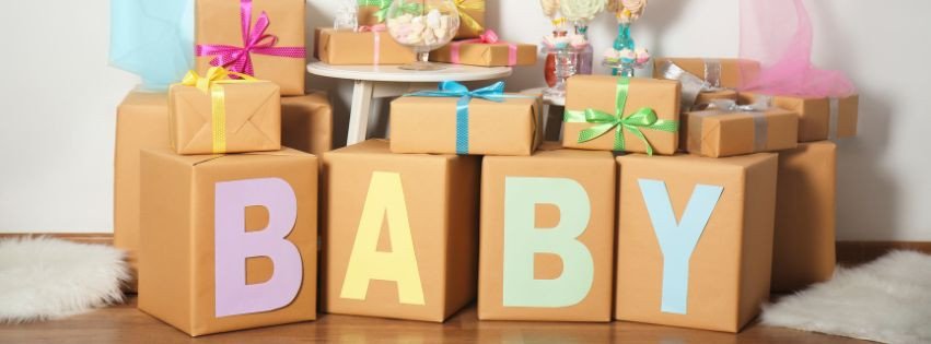 Cubes en carton formant le mot “BABY” avec des cadeaux emballés colorés et rubans, posé au sol dans une pièce. Propose le contexte “quoi offrir pour naissance bebe”.