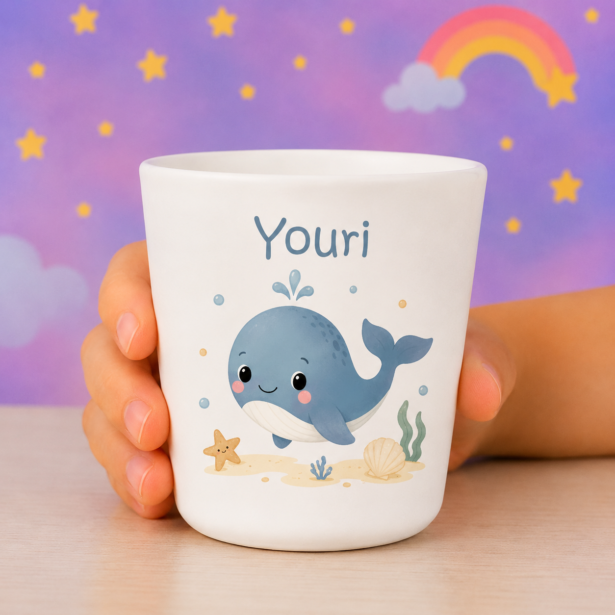 Gobelet réutilisable enfant personnalisé avec dessin de baleine bleu et texte Youri, tenu par une main, fond pastel avec étoiles et arc-en-ciel. gobelet réutilisable enfant avantages.