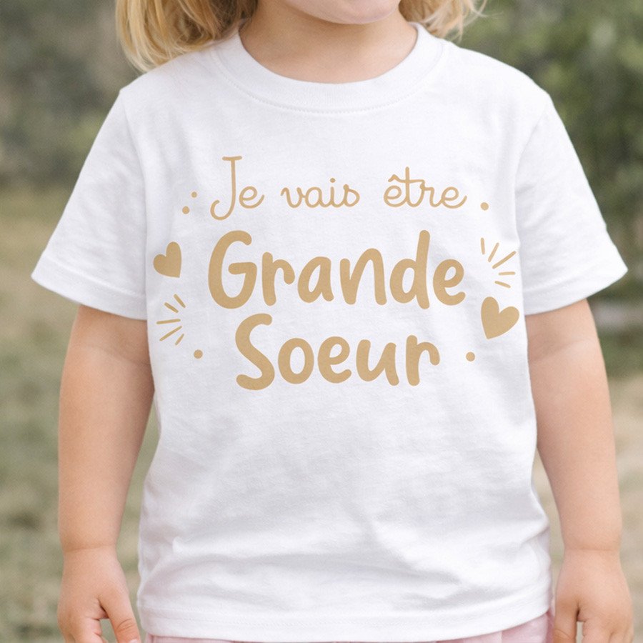 T-shirt blanc pour enfant avec le texte doré « Je vais être Grand e Sœur » décoratif et cœurs autour du message.