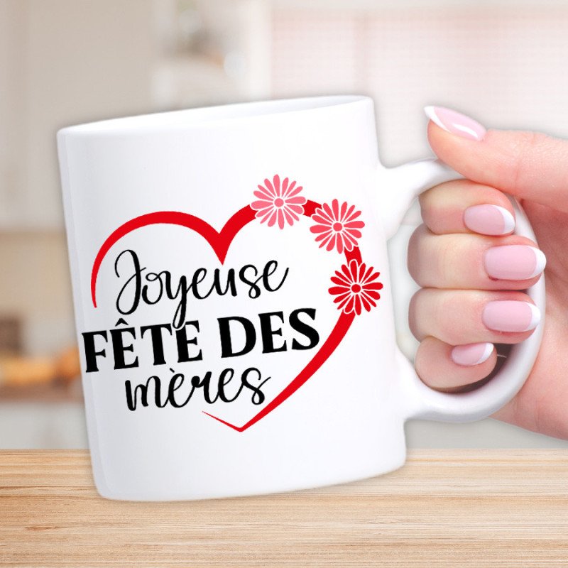 Tasse blanche avec texte « Joyeuse Fête des mères » entouré d’un cœur rouge et de fleurs roses, tenue par une main; idée de créer cadeau fête des mères personnalisé.